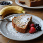 A slice of Chrissy Teigen's banana bread garnished with powdered sugar and berries.