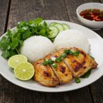 Served dish of grilled Vietnamese chicken with fresh herbs, lime wedges, rice, and dipping sauce, perfect for a savory meal.
