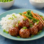 Delicious Teriyaki Chicken Meatballs served on a plate with sesame seeds and green onion garnish alongside rice and vegetables.