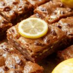 A close-up of vibrant lemon brownies arranged on a white serving platter. The brownies have a glossy lemon glaze on top, with a golden yellow hue and a fudgy texture visible on the edges. Soft, crumbly lemon zest is sprinkled over the surface, and the background features a rustic wooden table with fresh lemons nearby, enhancing the bright citrus theme.