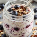 A clear glass jar filled with layered chia overnight oats topped with fresh blueberries and a drizzle of honey. The oats have a creamy texture with visible chia seeds, garnished with a sprig of mint. The jar is set on a rustic wooden surface with scattered blueberries and a small bowl of yogurt in the background.