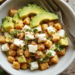 A vibrant bowl of Chickpea Feta Avocado Salad featuring creamy avocado slices, crisp chickpeas, and crumbled feta cheese, garnished with fresh herbs. The salad is arranged attractively in a white matte bowl, showcasing bright green avocado, golden chickpeas, and white feta contrasting against a light, colorful background with hints of cherry tomatoes and leafy greens.
