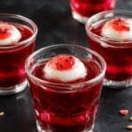 Close-up of eerie eyeball-shaped jello shots arranged on a dark platter. The gelatin is translucent with a red and white appearance resembling bloodshot eyes, complete with detailed irises and pupils. The textures of the gelatin are glossy and smooth, with some eyes slightly tilted to showcase their realistic design. Black edible decorations and dripping red syrup enhance the spooky effect, creating a chilling and festive display.