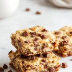 Close-up of a rectangular plate holding multiple no bake cookie dough bars, showcasing their golden-brown top with a slightly crumbly texture. The bars are topped with a drizzle of dark chocolate and sprinkled with chopped nuts, arranged neatly on a rustic wooden surface with a few scattered ingredients nearby.