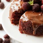 A luscious blackberry chocolate cake with a glossy dark chocolate glaze, topped with fresh blackberries and delicate chocolate shavings. The cake is layered, revealing rich chocolate and vibrant blackberries inside, set on an elegant white plate with a drizzle of berry sauce around it.
