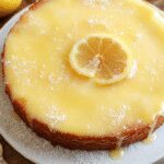 A slice of Lemon Custard Cake on a white plate, showcasing vibrant yellow lemon custard filling topped with a golden, slightly crispy crust. The cake's moist texture is visible, with a smooth, glossy custard layer contrasting with the fluffy cake edges. Fresh lemon slices and mint leaves garnish the plate for an appealing presentation.