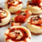 A close-up of three mini cheesecakes on a white plate. Each cheesecake has a smooth, creamy surface with a golden graham cracker crust visible at the base. Topped with fresh berries and a sprig of mint, they are neatly arranged with a soft, inviting appearance. The background features a rustic wooden table with scattered crumbs and a drizzle of berry sauce around the plate.