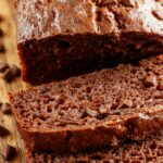 A rich, moist chocolate pumpkin bread loaf sliced open to reveal a dense, fudgy interior speckled with orange pumpkin pieces and glossy chocolate streaks, presented on a rustic wooden cutting board with a dusting of powdered sugar and a sprig of cinnamon for garnish.