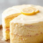 A beautifully layered lemon cake with a glossy lemon glaze dripping down the sides, topped with fresh lemon slices and delicate white flowers. The cake's moist texture is visible through its slices, and it rests on an elegant white cake stand, contrasting against a rustic wooden background.