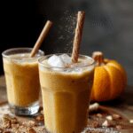 A tall glass of Pumpkin Chai Iced Latte with frothy milk on top, garnished with a cinnamon stick and a sprinkle of pumpkin spice, with a creamy beige and orange gradient from the latte, set against a rustic wooden table with fall-themed accents.