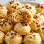 Close-up of round pumpkin cheesecake truffles coated in a smooth orange-colored coating, arranged on a rustic wooden platter with some crumbs and a cinnamon stick in the background. The truffles have a glossy finish, with a few partially bitten to reveal the creamy filling inside.