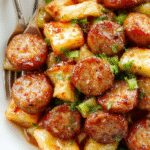 A vibrant skillet filled with golden-brown sausage slices, crunchy green cabbage, and colorful vegetables, all coated in a glossy glaze, garnished with fresh herbs, and served on a rustic wooden table with steam gently rising.