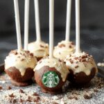 A close-up of a vibrant Starbucks cake pop coated in smooth, colorful icing, garnished with sprinkles, presented on a clean white plate with a blurred coffee cup in the background.