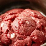 A close-up of vibrant red velvet cookie dough rolled into neat balls on a sleek white plate, with a few broken chunks revealing moist, crumbly texture, garnished with a sprinkle of white chocolate chips and a light dusting of cocoa powder, set against a rustic wooden background.