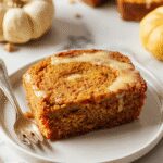 A slice of pumpkin bread topped with a swirl of creamy vanilla pudding and a dusting of cinnamon. The bread is golden-brown with a moist interior and visible chunks of pumpkin, presented on a rustic wooden plate with a background of fall-themed decor and a spoon resting nearby.