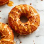 A close-up of baked pumpkin donuts arranged on a rustic wooden platter. The donuts are golden-brown with a dusting of cinnamon and pumpkin spice. A glossy maple glaze drips slightly from some of the donuts, highlighting their sticky, sweet coating. The presentation includes a small bowl of extra maple glaze and scattered cinnamon sticks and pumpkin seeds in the background, creating a warm, autumnal atmosphere.
