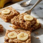 Golden-brown banana oatmeal bars arranged on a rustic wooden surface, showing a slightly crumbly texture with visible banana slices and oats, some bars cut open to reveal a moist interior with chunks of banana and oats, garnished with a few banana slices and oat flakes.