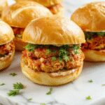 A close-up of three golden-brown chicken sliders arranged on a white platter, topped with creamy bang bang sauce, garnished with chopped green onions, and served alongside fresh salad greens, all styled against a rustic wooden background.