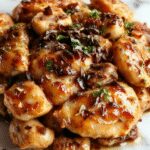 A glossy stir-fry dish featuring tender chicken pieces coated in a dark black pepper sauce, surrounded by sliced mushrooms and vibrant green garnishes, served on a chic white plate with sesame seeds sprinkled on top, garnished with chopped scallions.