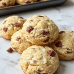 A close-up of golden-brown protein biscuits arranged on a rustic wooden plate, with visible textured surface and a few biscuits slightly broken to reveal a moist, dense interior. The background features a softly blurred breakfast setting with a coffee cup and fresh fruit.
