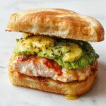 A close-up of a toasted sandwich with golden-brown bread crust, filled with creamy sliced avocado, tender cooked chicken, and melted cheese. The sandwich is cut in half, revealing the colorful layers inside, garnished with fresh herbs on a rustic wooden board.