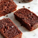 A golden-brown loaf of chocolate espresso banana bread sits on a rustic wooden surface. The top is sprinkled with dark chocolate chips and a light dusting of powdered sugar, with slices slightly fanned out revealing a moist, dense interior flecked with chocolate chunks and banana pieces. The warm tones and inviting texture make it look both decadent and comforting.