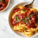 A close-up of a rustic white plate filled with vibrant, steaming spaghetti coated in rich, crimson tomato sauce garnished with fresh basil leaves, surrounded by various ingredients like garlic cloves, olive oil, and grated cheese on a wooden table.