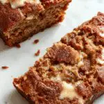 A slice of moist banana bread topped with a swirl of butter on a rustic wooden plate, with banana slices and a sprig of mint beside it, spotlighted with soft natural light