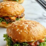 A close-up of a juicy grilled burger patty nestled in a toasted sesame seed bun topped with fresh lettuce, ripe tomato slices, melted cheese, and pickles, with a side of crispy golden fries on a rustic wooden plate.