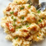 A close-up of a creamy, golden-brown chicken breast smothered in rich sauce, served atop a bed of fluffy rice with garnishes of fresh herbs and vegetables, set on a rustic plate with a vibrant background.