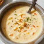 A rustic white bowl filled with creamy crack potato soup topped with shredded cheese, chopped green onions, and bacon bits. The soup has a thick, velvety texture with visible chunks of potatoes and melted cheese, presented on a wooden table with a spoon resting beside it. In the background, a slow cooker is partially visible, hinting at the comforting, homemade nature of the dish.