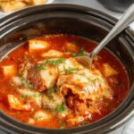 A steaming bowl of lasagna soup with layers of rich tomato broth, melted cheese, and tender lasagna noodles topped with fresh basil and grated Parmesan, served on a rustic wooden table with a spoon resting beside it.