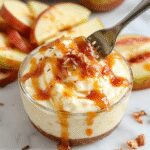 A close-up of a creamy caramel apple cheesecake dip in a clear glass bowl, topped with chunks of fresh green and red apples, drizzled with golden caramel sauce, surrounded by apple slices and crunchy graham cracker crumbs, all set on a rustic wooden table with fall-themed decor in the background.