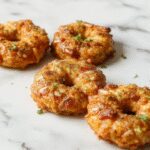 Golden-brown apple fritters glazed with a shiny sugar coating, crispy on the outside with visible apple pieces inside, arranged on a white plate garnished with cinnamon sticks and fresh apple slices, set against a rustic wooden background.