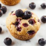 A close-up of a golden-brown blueberry protein muffin topped with fresh blueberries and a light dusting of powdered sugar, placed on a rustic wooden plate with a few blueberries scattered around. The muffin's texture is moist and fluffy, with visible bursts of juicy blueberries and a slightly cracked crust revealing its soft interior.