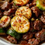 A vibrant zucchini beef stir-fry served in a white bowl, featuring thin slices of beef and julienned zucchini coated in a glossy sauce, garnished with chopped scallions and sesame seeds, with colorful vegetables in the background.