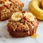 A golden-brown loaf of honey vanilla banana bread placed on a rustic wooden surface, sliced to reveal a moist, fluffy interior dotted with ripe banana chunks, topped with a drizzle of honey and a sprinkle of vanilla bean specks, with fresh bananas and a small jar of honey nearby.