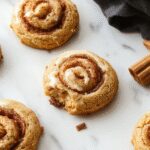 A close-up of golden-brown cinnamon roll cookies arranged on a rustic wooden platter. The cookies are swirled with cinnamon sugar and glisten slightly, showcasing their soft, flaky texture. Some cookies are topped with a light dusting of powdered sugar, with a few broken pieces revealing their tender interior. The background features a cozy setting with a warm cup of coffee and cinnamon sticks, enhancing the inviting atmosphere.