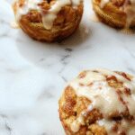 A close-up of golden-brown peach pie cruffins arranged in a circular pattern on a white plate, showcasing flaky crust, juicy peach filling peeking through the layers, and a dusting of powdered sugar on top.