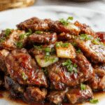A sizzling Chinese Pepper Steak with onions is presented on a white ceramic plate. The tender beef strips coated in a glossy, dark soy-based sauce are topped with crisp, caramelized onion slices and sprinkled with freshly ground black pepper. The dish is garnished with sliced green onions and sesame seeds, set against a dark wooden surface with a few scattered peppercorns around it. The vibrant colors of the beef, onions, and green garnishes beautifully contrast with the shiny sauce and rustic plate.