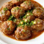 A close-up of a rustic white bowl filled with golden-brown Salisbury steak meatballs in a rich gravy, garnished with chopped parsley. The meatballs are tender and juicy, surrounded by a glossy, savory sauce, with some meatballs partially submerged to showcase their texture. The background features a blurred cozy kitchen setting with warm tones.