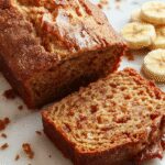 A golden-brown loaf of snickerdoodle banana bread sits on a rustic wooden table, topped with a cinnamon-sugar swirl and a light dusting of cinnamon. The breadβs surface shows cracks and a tempting crust, with slices revealing a moist, banana-filled crumb inside. The background includes a cinnamon stick and a small bowl of cinnamon-sugar mixture, enhancing the warm, cozy feel.