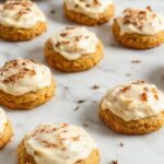 A close-up of two soft pumpkin cookies topped with creamy cinnamon frosting, beautifully arranged on a rustic wooden plate, with a dusting of cinnamon around them. The cookies are golden orange with a slightly cracked surface, showcasing their moist texture and warm spice notes.