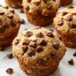 A close-up of a golden-brown banana chocolate chip muffin with a slightly cracked top, dotted with melted chocolate chips, placed on a rustic wooden surface with a few sliced bananas and chocolate chips scattered nearby. The muffinβs texture looks moist and tender with visible banana chunks and gooey chocolate inclusion.