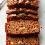 A golden-brown zucchini bread loaf sliced to reveal a moist, green-speckled interior with a slightly cracked top, presented on a rustic wooden board with a few green zucchini beside it. The slices show a tender crumb, with visible bits of shredded zucchini throughout, emphasizing its moist texture and inviting appearance.