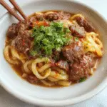 A bowl of beef and noodles garnished with chopped green onions and sesame seeds. The dish features tender ground beef mixed with al dente pasta and a glossy sauce, presented on a rustic wooden table with scattered herbs and vegetables in the background.