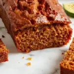 A golden-brown loaf of zucchini pumpkin bread topped with a light dusting of powdered sugar, sliced to reveal moist, speckled interior with hints of orange and green, presented on a rustic wooden board with fresh zucchini and pumpkin placed nearby.