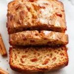 A warm, freshly baked loaf of apple cinnamon bread sits on a rustic wooden surface, sliced open to reveal a fragrant, spiced apple filling. The bread has a golden-brown crust with a textured, slightly cracked top, topped with a dusting of powdered sugar. Sliced pieces are fanned out around the loaf, showcasing the moist, swirled interior filled with cinnamon and apple chunks. The background features a cozy fall setting with autumn leaves and a mug of steaming coffee or tea nearby.