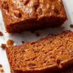 A slices of Chai Spice Maple Pumpkin Bread on a rustic wooden platter, topped with a light dusting of powdered sugar, with a steaming cup of chai tea in the background. The bread has a golden-brown crust with swirls of spiced pumpkin filling visible inside, garnished with a cinnamon stick and a sprinkle of pumpkin seeds for texture.