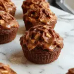 A tray of rich chocolate pumpkin cupcakes topped with swirls of creamy orange frosting, decorated with tiny chocolate chips and a subtle dusting of cinnamon, presented on a rustic wooden surface with fall leaves and mini pumpkins in the background.